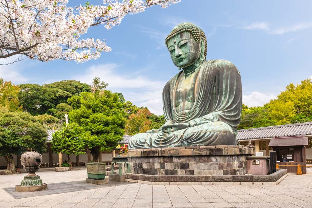 Il Grande Buddha di Kamakura, un'enorme statua di bronzo dalla patina verde, seduto all'aperto su un basamento di pietra nel cortile del tempio Kotoku-in. In alto a sinistra spuntano i rami di un ciliegio in fiore, e sullo sfondo si vedono alberi verdi ed edifici tradizionali sotto un cielo azzurro