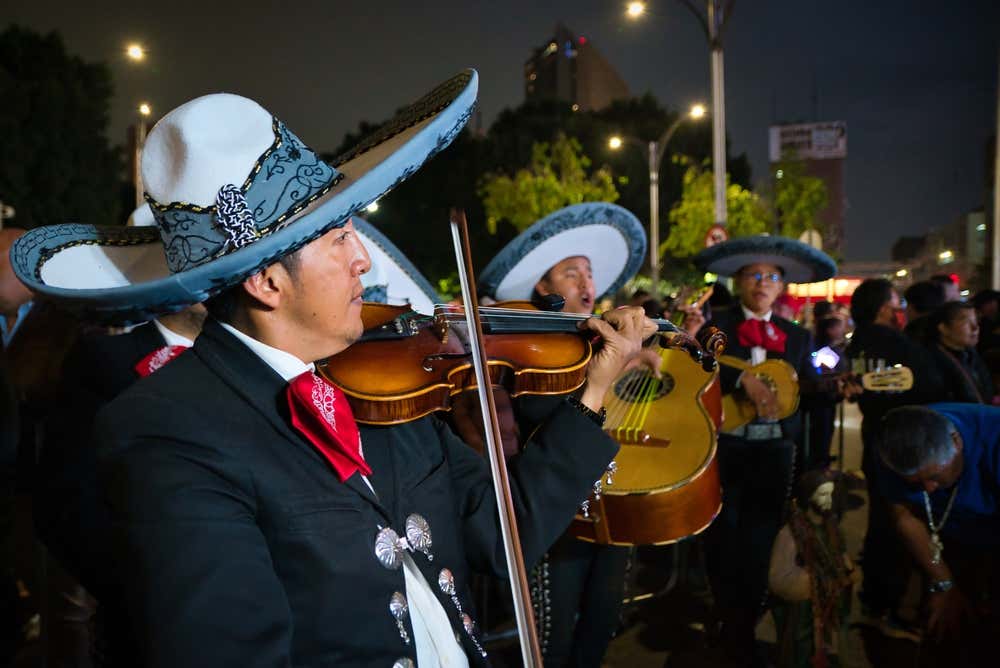 A mariachi band performing with violins, guitars and traditional clothing at night.