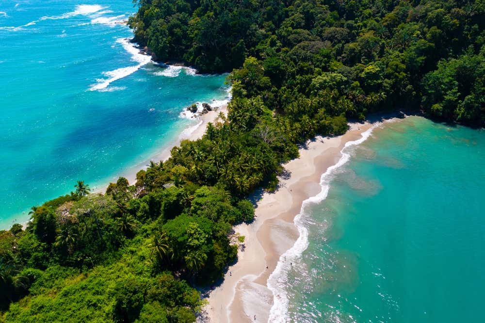 Playa Manuel Antonio seen from above with the water visible on either side of a beach with trees in the middle.