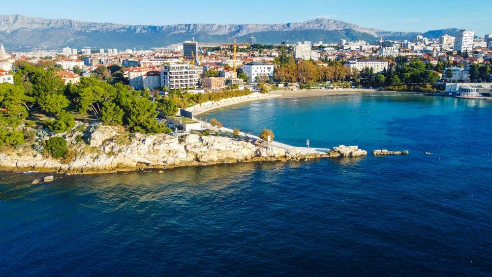 Panorama aereo della baia cittadina di Bacvice a Spalato, con la sua spiaggia sabbiosa a mezzaluna, il mare turchese, le pinete costiere e gli edifici della città sormontati dalle montagne sullo sfondo