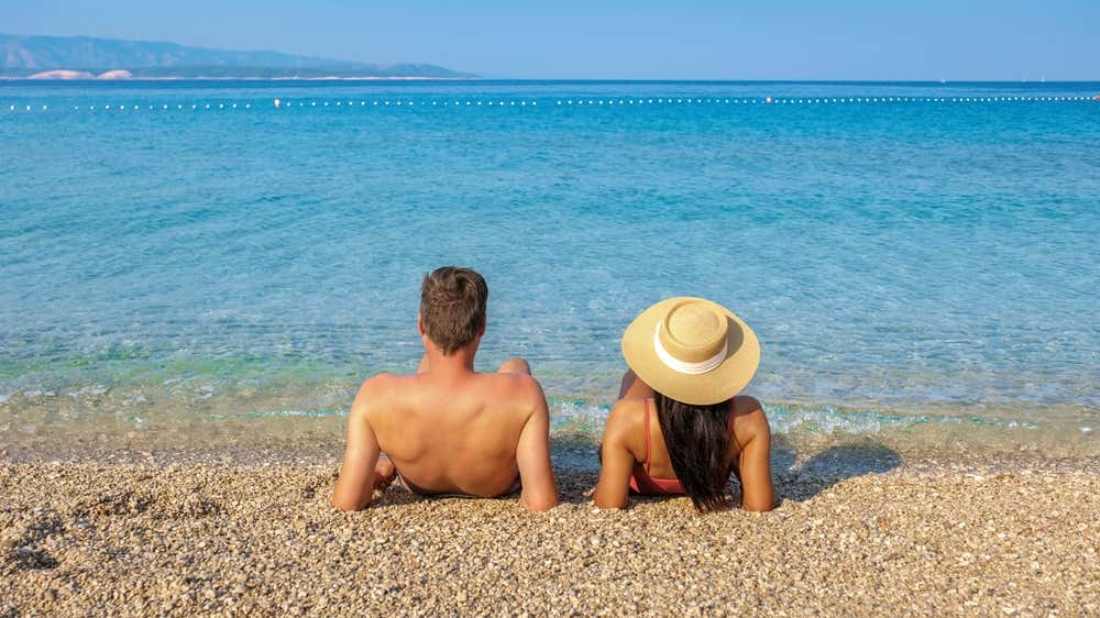 Una coppia vista di spalle sdraiata al sole sulla spiaggia di ciottoli di Zlatni Rat (Corno d'Oro) nell'isola di Brač, di fronte a un mare turchese e cristallino