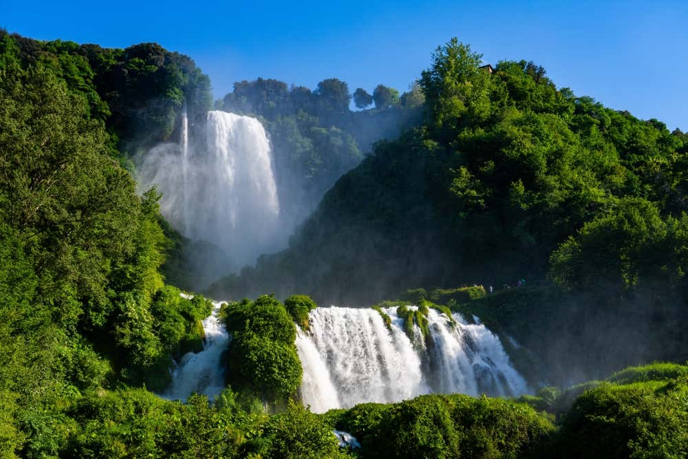 La Cascata delle Marmore, vicino a Terni, in piena portata: l'acqua bianca e spumeggiante scende in più salti tra pareti di roccia coperte da una fitta vegetazione verde. Tutto intorno si alza una nuvola di spruzzi, sotto un cielo azzurro limpido