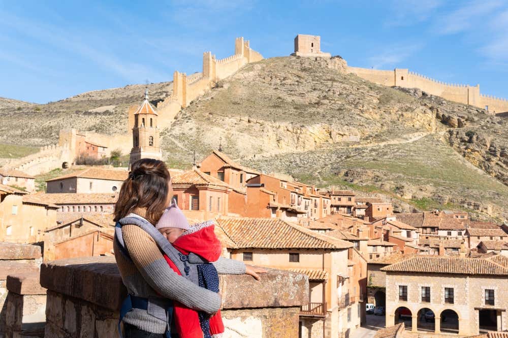 Una madre con il suo neonato nel marsupio guarda il panorama del borgo medievale di Albarracín, con le sue tipiche case dai toni ocra e le imponenti mura difensive che risalgono la collina sullo sfondo