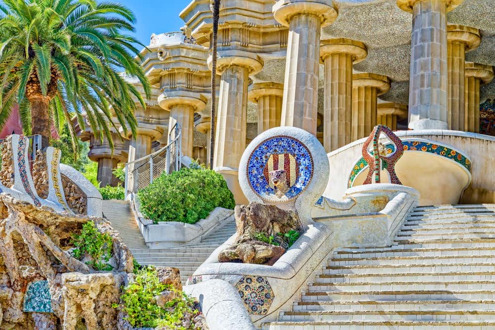 La scalinata monumentale del Parc Güell a Barcellona, con i parapetti decorati a trencadís e un medaglione a mosaico con lo stemma catalano e una testa di serpente. In cima alle scale si vedono le colonne doriche della Sala Ipostila, affiancate da una palma e da vegetazione mediterranea