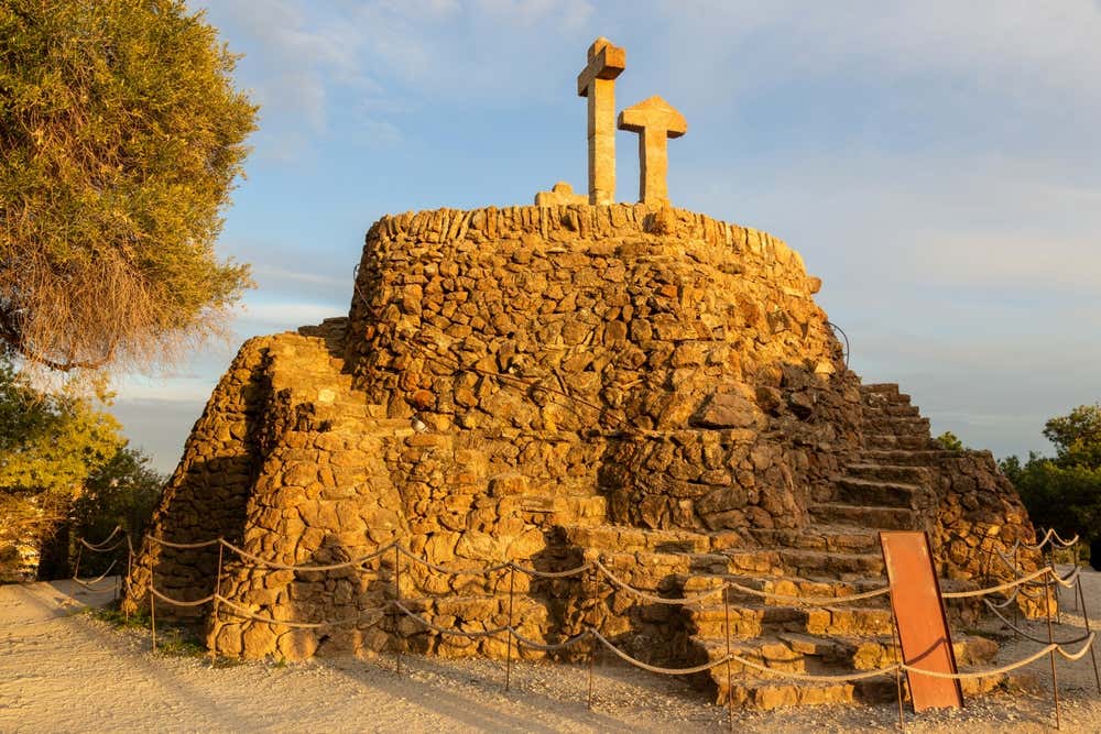 Il Calvario del Parc Güell a Barcellona, detto anche Turó de les Tres Creus: un tumulo di pietre grezze con una scalinata che sale fino alle croci in pietra sulla cima. Il tumulo è illuminato dalla luce dorata del tramonto e circondato da una staccionata di corda