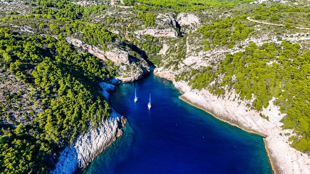 Veduta aerea dall'alto della baia di Stiniva sull'isola di Vis, con alte scogliere rocciose ricoperte di vegetazione che racchiudono un'insenatura dal mare blu intenso in cui sono ancorate due barche a vela