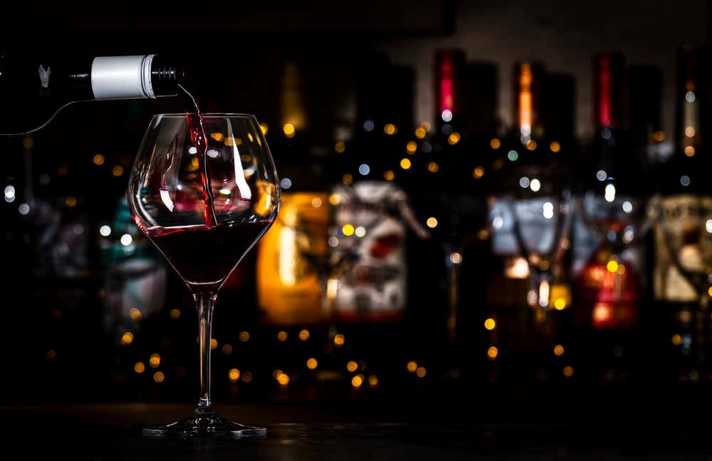Una copa de cristal en la que se vierte vino tinto de calidad, con un fondo oscuro iluminado por luces cálidas tenues y botellas de vinoteca