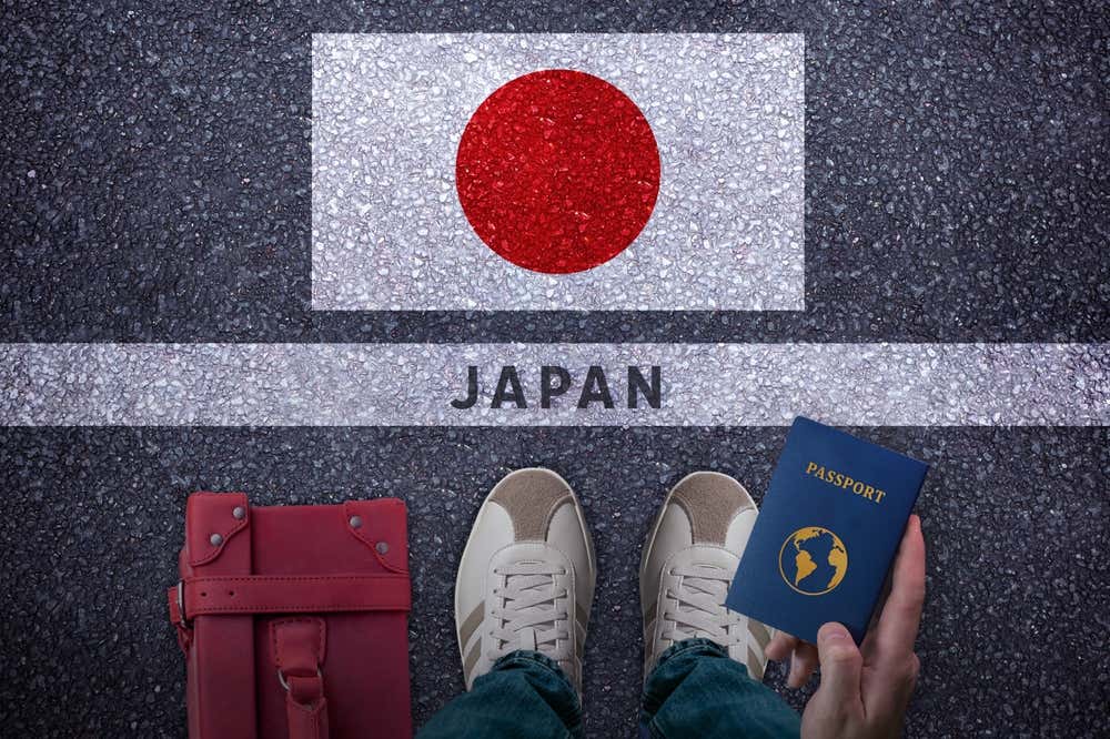 Una persona vista dall'alto, in piedi sull'asfalto davanti alla bandiera del Giappone dipinta a terra, con la scritta "Japan". Tiene in mano un passaporto blu e accanto ai suoi piedi c'è una borsa da viaggio rossa