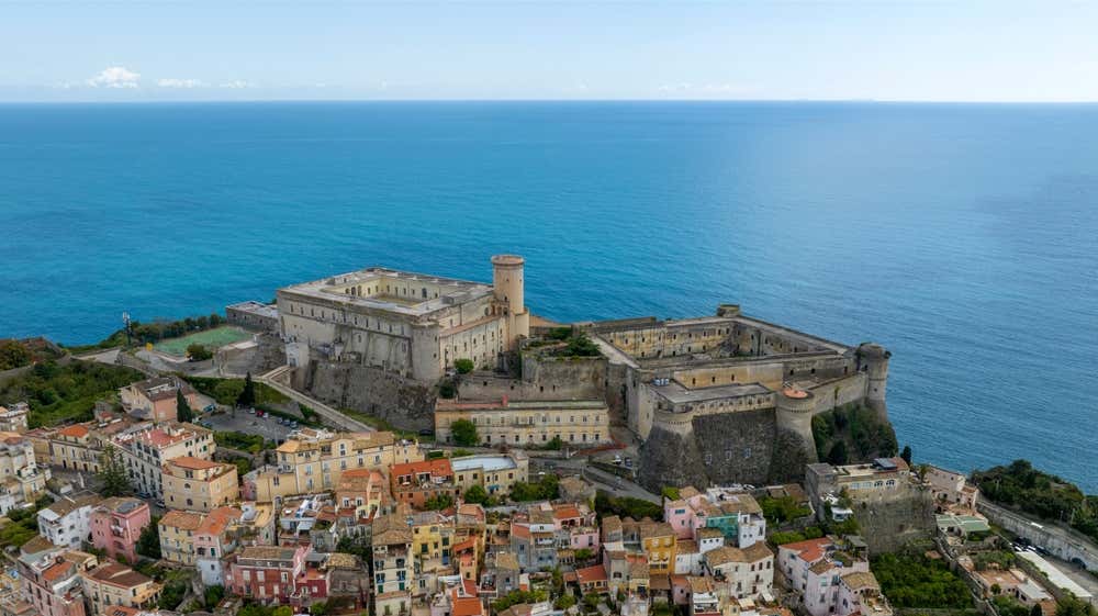 Vista aerea di Gaeta, sulla costa del Lazio meridionale. Sul promontorio si trova il Castello angioino-aragonese, con torri e mura in pietra affacciate sul mare. In primo piano ci sono le case colorate del centro storico e sullo sfondo il mare aperto, di un blu intenso