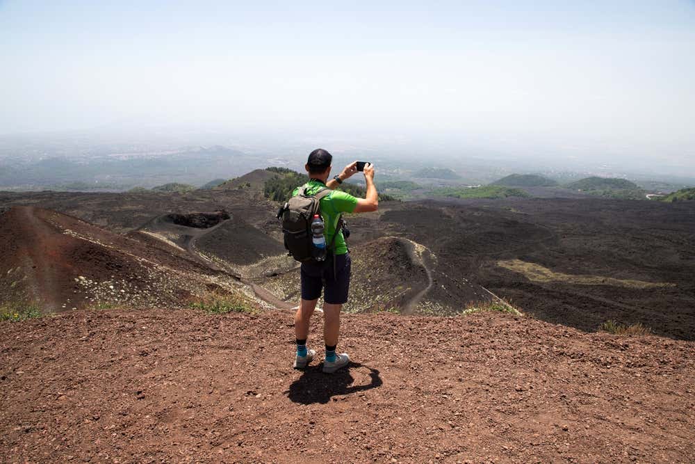 Un escursionista con zaino scatta una foto con lo smartphone sui crateri e i paesaggi vulcanici dell'Etna, circondato da rocce scure e terra rossa