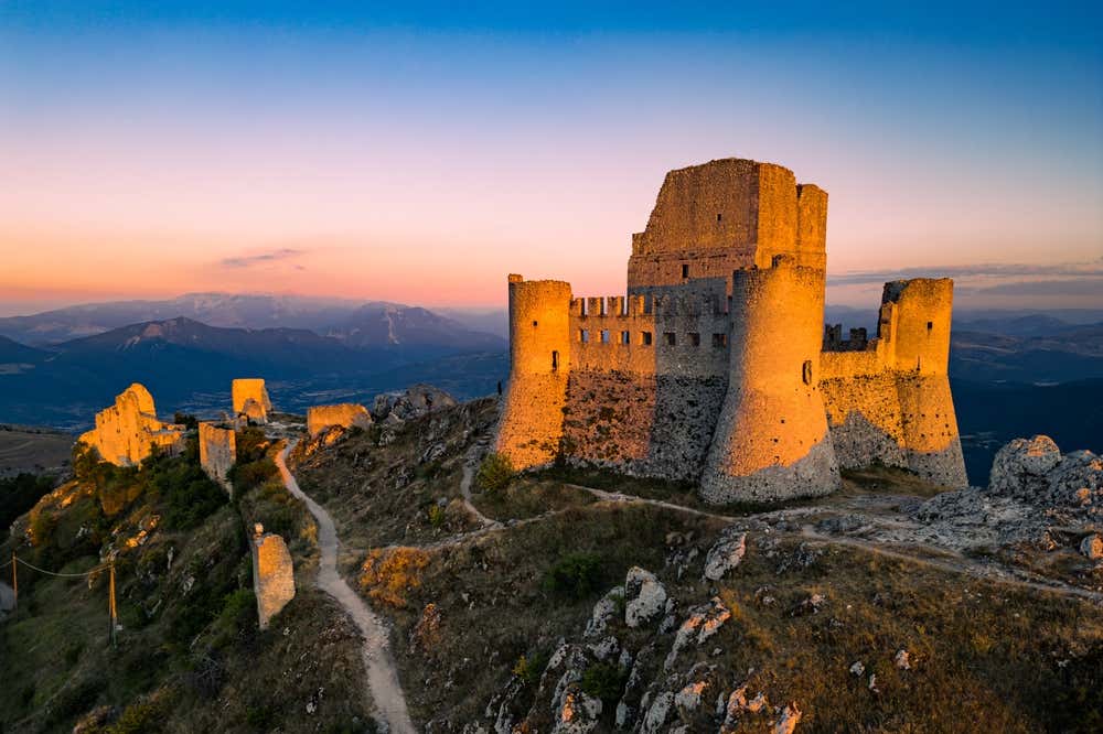 Rocca Calascio al tramonto, in Abruzzo: una fortezza medievale in pietra con torri circolari e un mastio centrale, sulla cima di un crinale roccioso. Accanto si vedono i ruderi del borgo e un sentiero che sale verso il castello, mentre sullo sfondo le montagne sfumano sotto un cielo tra il rosa e l'azzurro