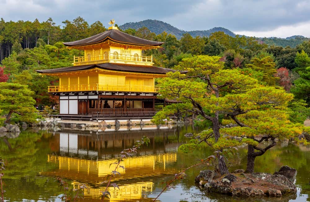 Il Kinkaku-ji, il Padiglione d'oro di Kyoto: un tempio zen a tre piani, con i due superiori ricoperti di foglia d'oro e una fenice dorata sul tetto. Il tempio si riflette nelle acque di uno stagno. In primo piano c'è un pino potato con cura su un isolotto di roccia, mentre sullo sfondo si vedono colline boscose con qualche tocco rosso autunnale