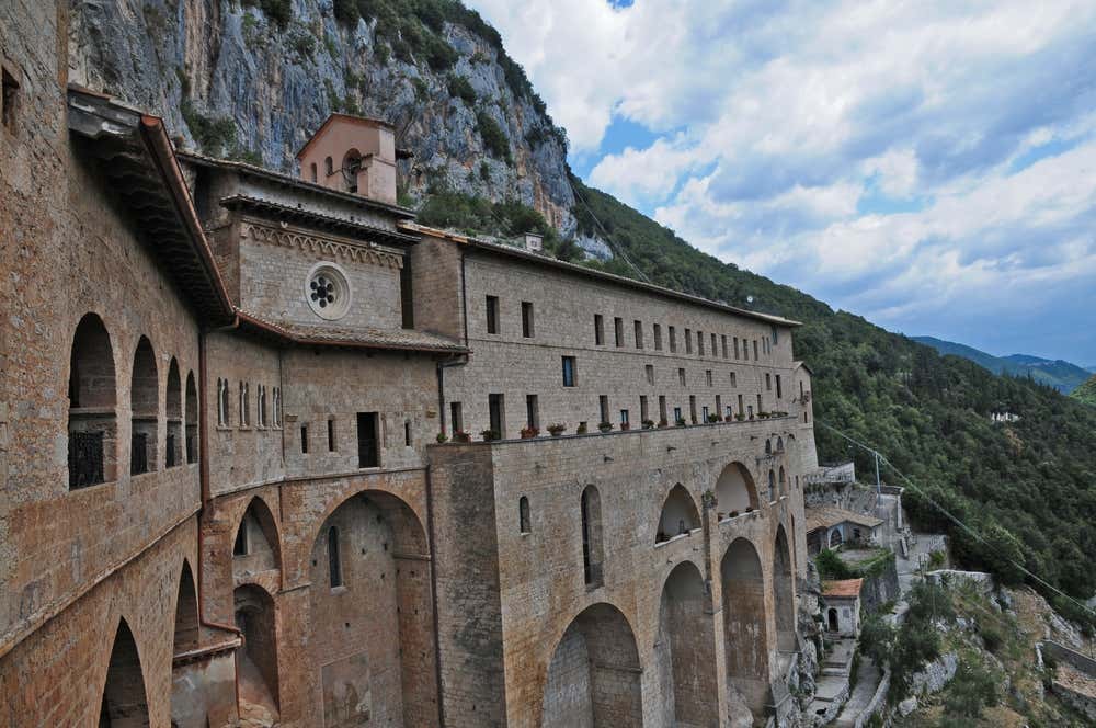 Il monastero di San Benedetto, detto Sacro Speco, a Subiaco: un complesso in pietra con archi alti, un campanile e un rosone, costruito a ridosso di una parete di roccia. Intorno si vedono montagne coperte di boschi nella valle dell'Aniene, sotto un cielo con nuvole bianche
