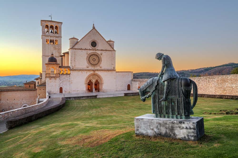 La facciata della Basilica superiore di San Francesco ad Assisi, in pietra chiara con un grande rosone e il campanile, al tramonto. In primo piano, su un prato verde, c'è una statua in bronzo di San Francesco a cavallo, con il capo chino. Sullo sfondo si vedono le colline umbre sotto un cielo sfumato d'arancio