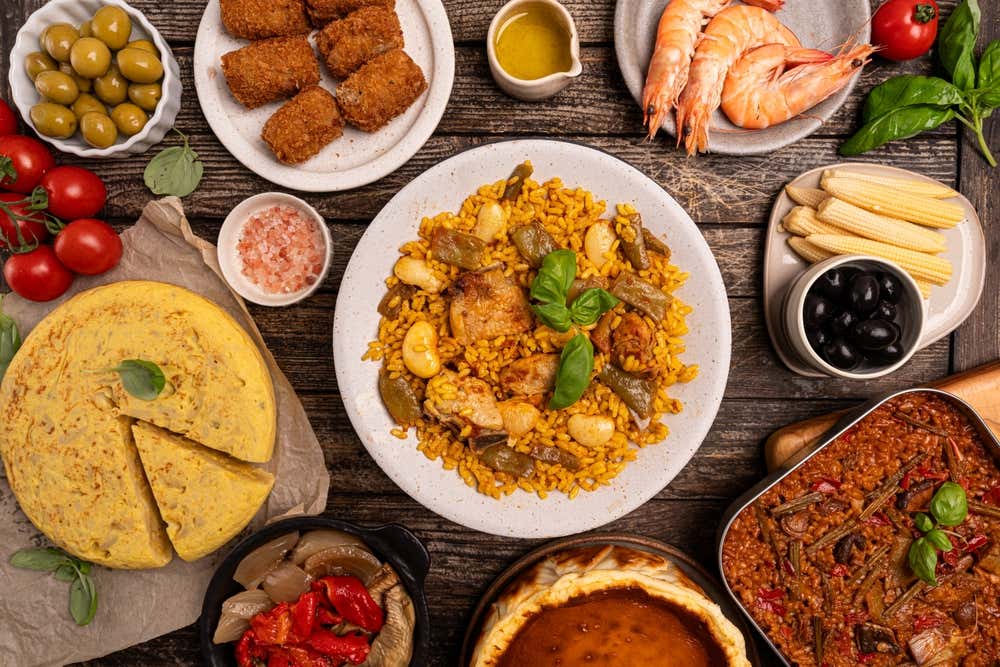 Vista dall'alto di un tavolo in legno ricco di piatti tipici della cucina spagnola, tra cui una paella valenciana al centro, una tortilla de patatas, crocchette, gamberi, olive e tapas varie
