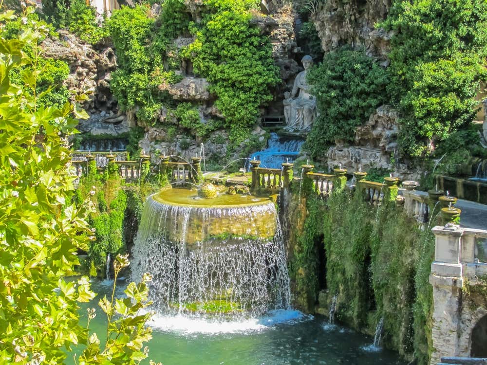 La Fontana dell'Ovato a Villa d'Este, a Tivoli: l'acqua scende a cascata da una vasca ovale in una piscina verde smeraldo. Tutto intorno ci sono balaustre in pietra, rocce ricoperte di vegetazione e, sullo sfondo, una statua femminile seduta in una grotta