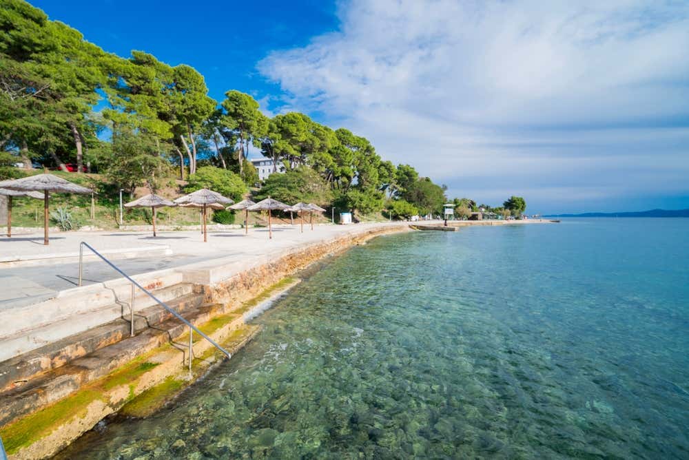 Una tranquilla passeggiata sul mare pavimentata con gradini in pietra per accedere alle acque limpide e turchesi, affiancata da ombrelloni in paglia e una fitta pineta sotto un cielo azzurro in Croazia
