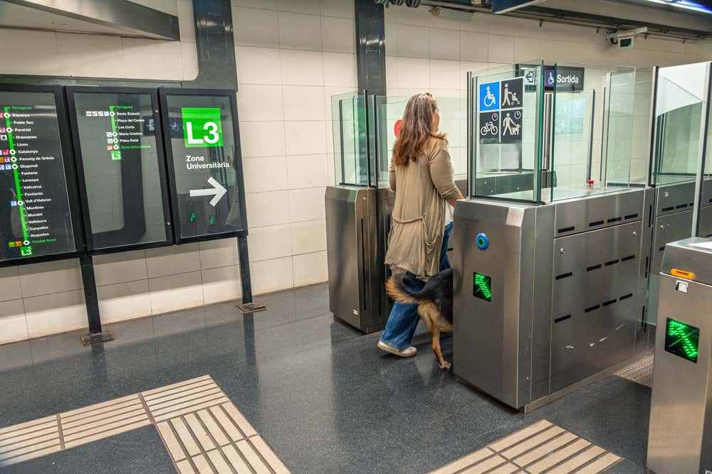 Una donna con un cane passa i tornelli di una stazione della metropolitana di Barcellona. Sulla sinistra c'è un pannello verde della linea L3 in direzione Zona Universitària, con la mappa delle fermate. In alto si legge l'indicazione "Sortida", cioè uscita