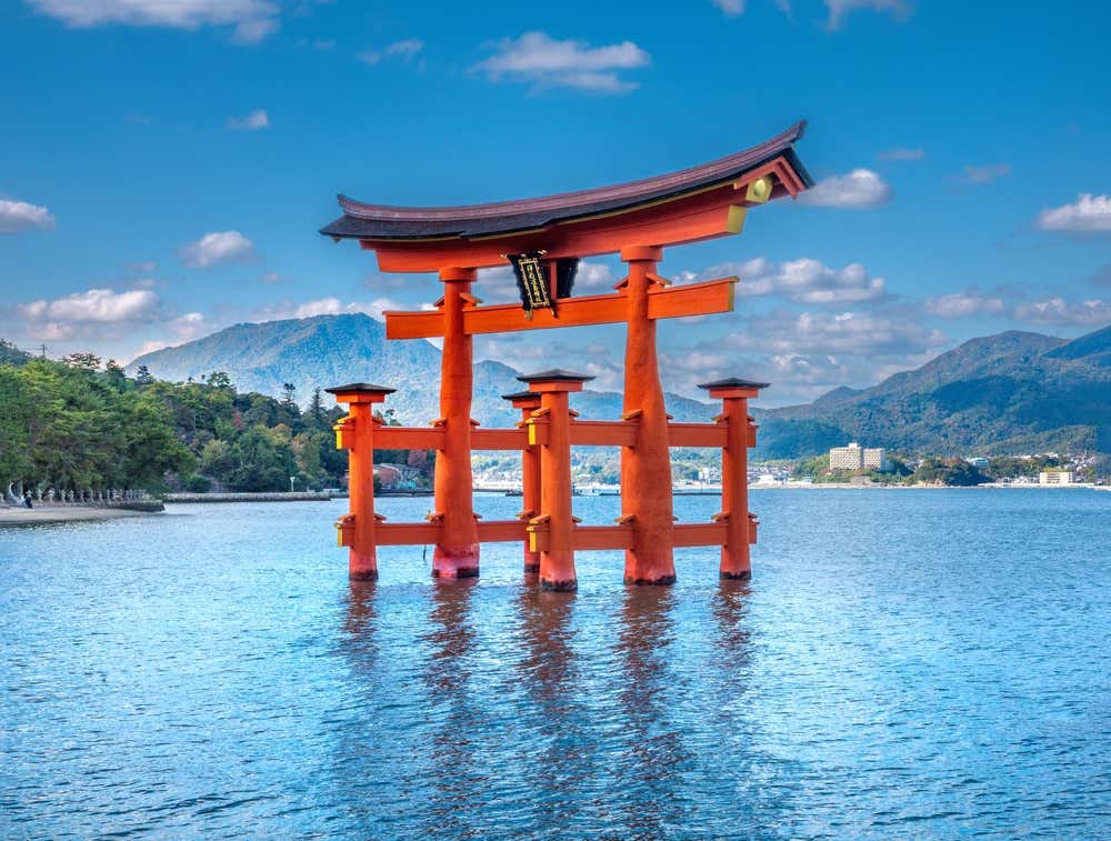 Il grande torii arancione del santuario di Itsukushima, sull'isola di Miyajima, emerge dal mare durante l'alta marea e si riflette nell'acqua. Sullo sfondo ci sono montagne verdi, la costa e un cielo azzurro con qualche nuvola bianca