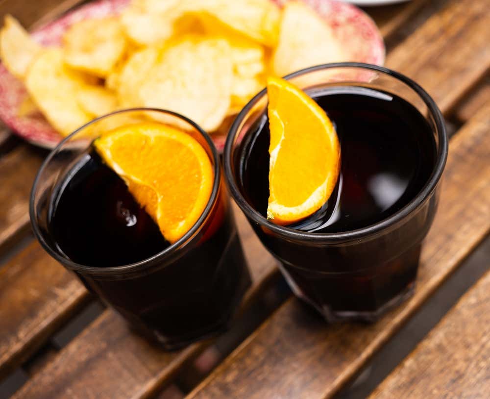 Dos copas de vermut rojo servidas con una rodaja de naranja sobre una mesa de madera, con un platito de patatas fritas de fondo para el aperitivo