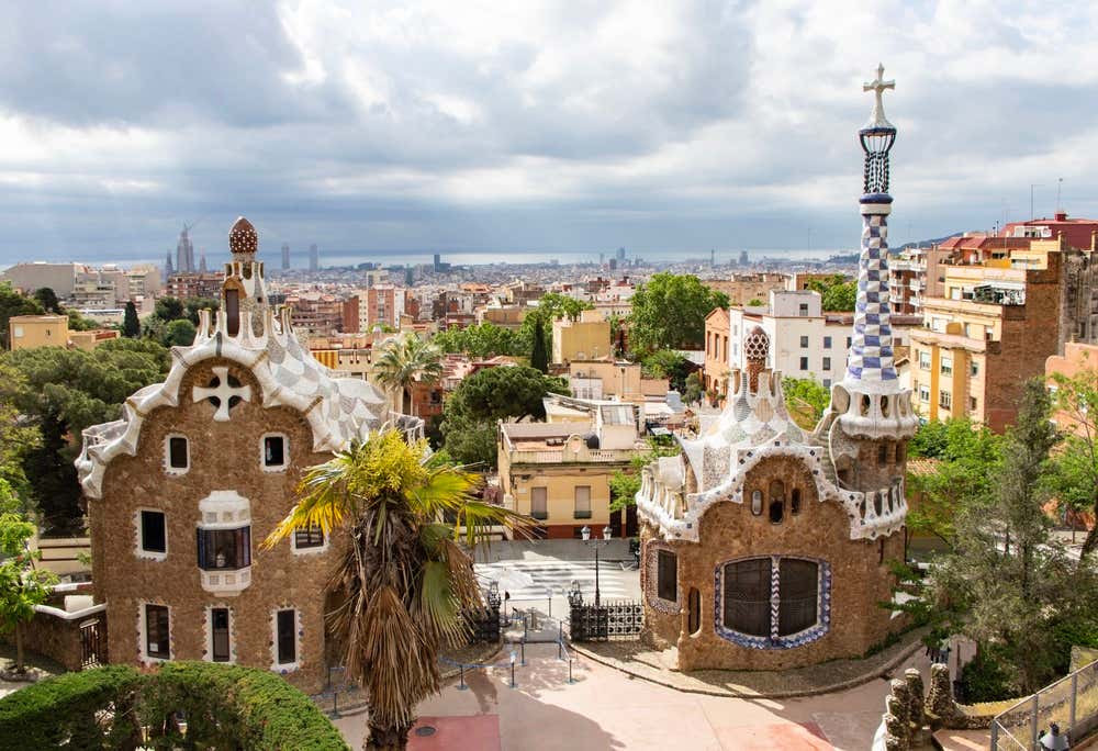 I due padiglioni d'ingresso del Parc Güell a Barcellona: hanno i tetti ricoperti di trencadís bianco e una torre a scacchi bianchi e blu con in cima una croce. Sullo sfondo si vede la città fino al mare, sotto un cielo nuvoloso