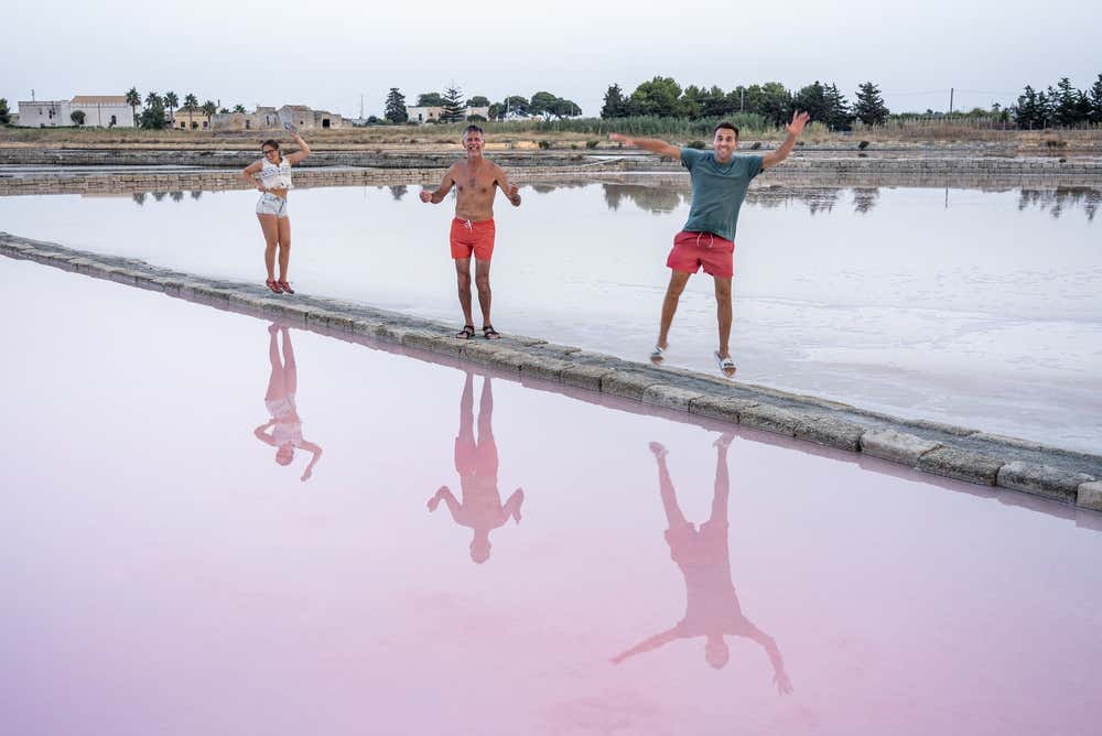 Tre persone felici (una donna e due uomini) posano in piedi sull'argine di una vasca rosa delle Saline di Trapani, con i loro riflessi nitidi specchiati nell'acqua
