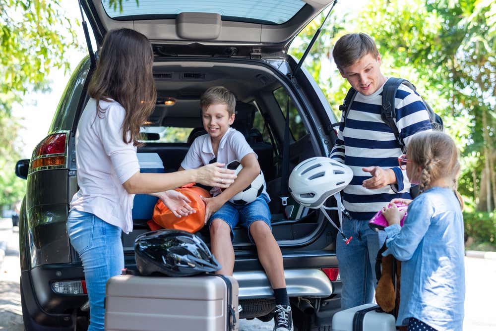 Una famiglia con due bambini carica il bagagliaio di un'auto prima di partire. Un bambino, seduto nel portabagagli, tiene in mano un pallone da calcio, mentre la bambina stringe un borsello rosa. I genitori sistemano borse, caschi da bici e valigie, all'ombra degli alberi in una giornata di sole