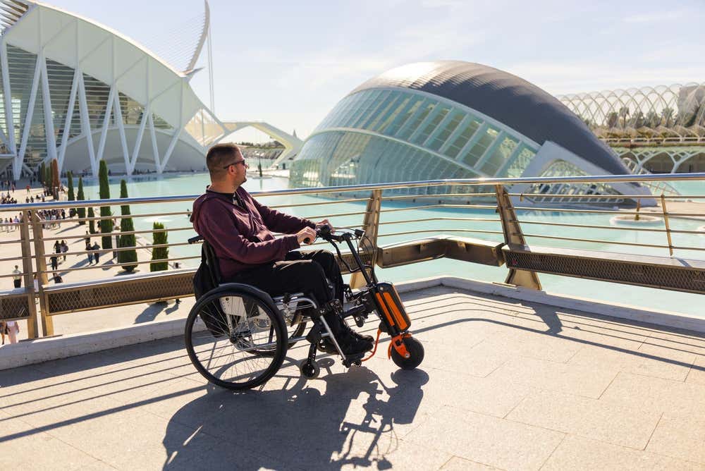 Un uomo su una sedia a rotelle motorizzata ammira il panorama futuristico della Città delle Arti e delle Scienze di Valencia, con le sue suggestive strutture bianche come L'Hemisfèric riflesse nelle vasche d'acqua