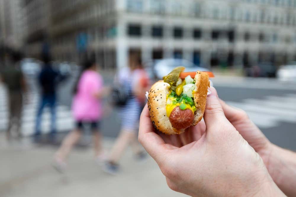 Someone holding a hot dog with a sesame seed bun,pickles, onions, peppers and other garnishes.
