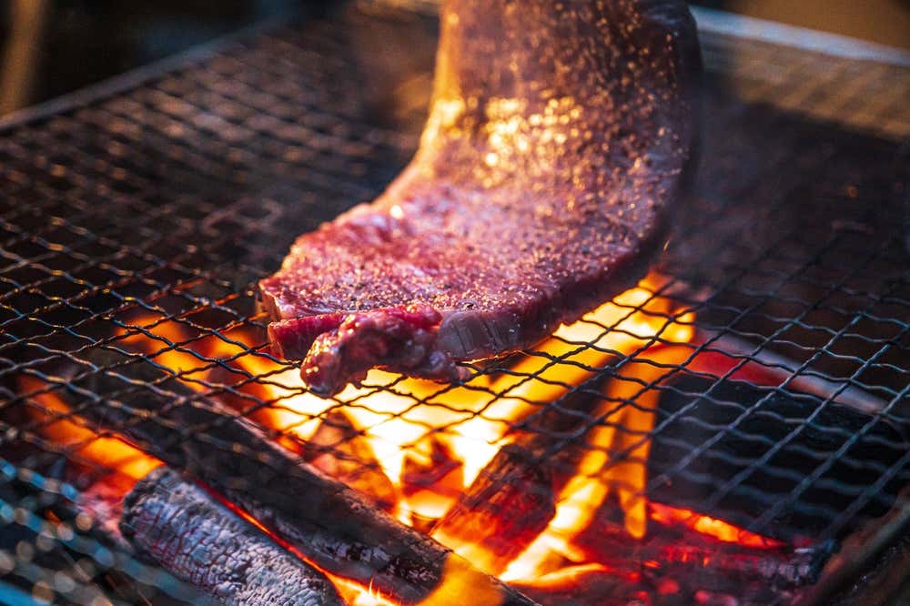 A raw steak being placed onto a grill with an orange fire burning on wood below.