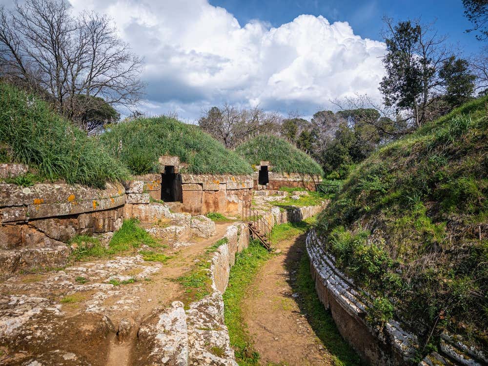 La necropoli etrusca della Banditaccia a Cerveteri: tumuli circolari in tufo coperti d'erba, con gli ingressi scuri delle tombe e sentieri di terra battuta che passano tra le pareti di roccia. Sullo sfondo ci sono alberi e grandi nuvole bianche in un cielo azzurro