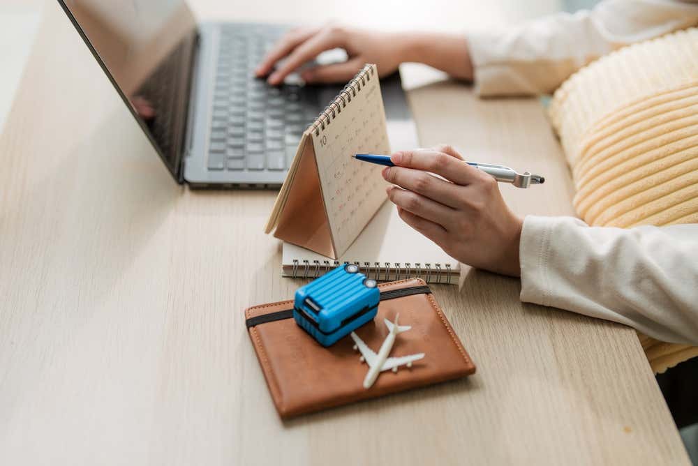 Una persona pianifica un viaggio alla scrivania: con una mano scrive al computer portatile e con l'altra indica con la penna una data su un calendario da tavolo. In primo piano, su un portadocumenti in pelle marrone, ci sono una piccola valigia azzurra e un aeroplanino giocattolo