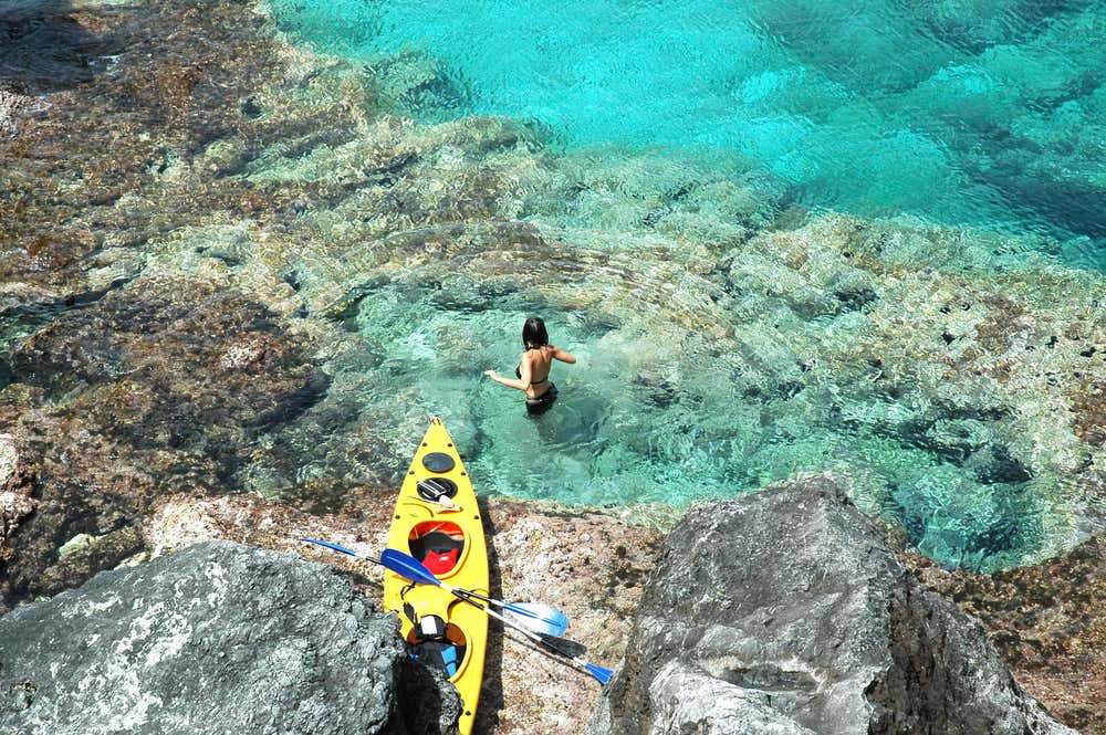 Una donna in piedi nell'acqua cristallina color turchese delle Isole Tremiti, accanto a un kayak giallo parcheggiato su una costa rocciosa. Scatto dall'alto