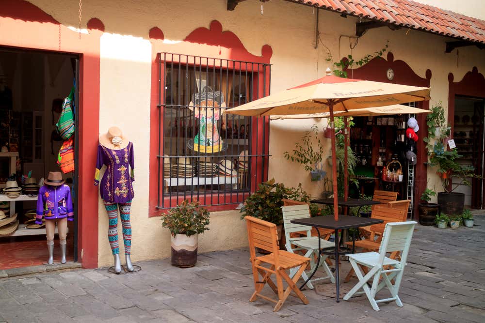 A street in Tlaquepaque with a hat shop on the left side and a couple of tables with parasols and chairs.