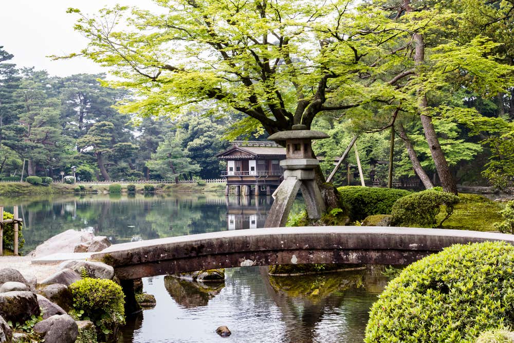 Il giardino Kenroku-en di Kanazawa, con la celebre lanterna in pietra Kotoji-toro a due gambe accanto a uno stagno. In primo piano c'è un ponticello di pietra ad arco. Sullo sfondo si vede una casa da tè in legno su palafitte che si riflette nell'acqua, sotto alberi dal fogliame verde chiaro