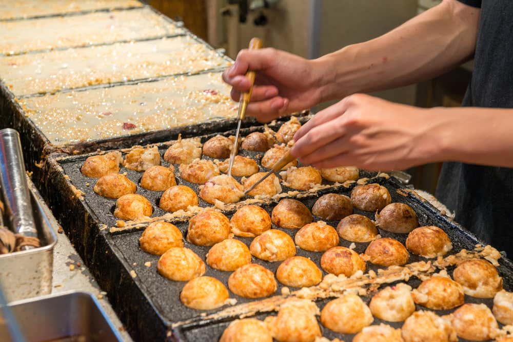 Un cuoco prepara i takoyaki su una piastra in ghisa con tanti incavi rotondi. Con due bastoncini appuntiti gira le polpette di pastella dorate, mentre sullo sfondo si vedono altre piastre con la pastella appena versata