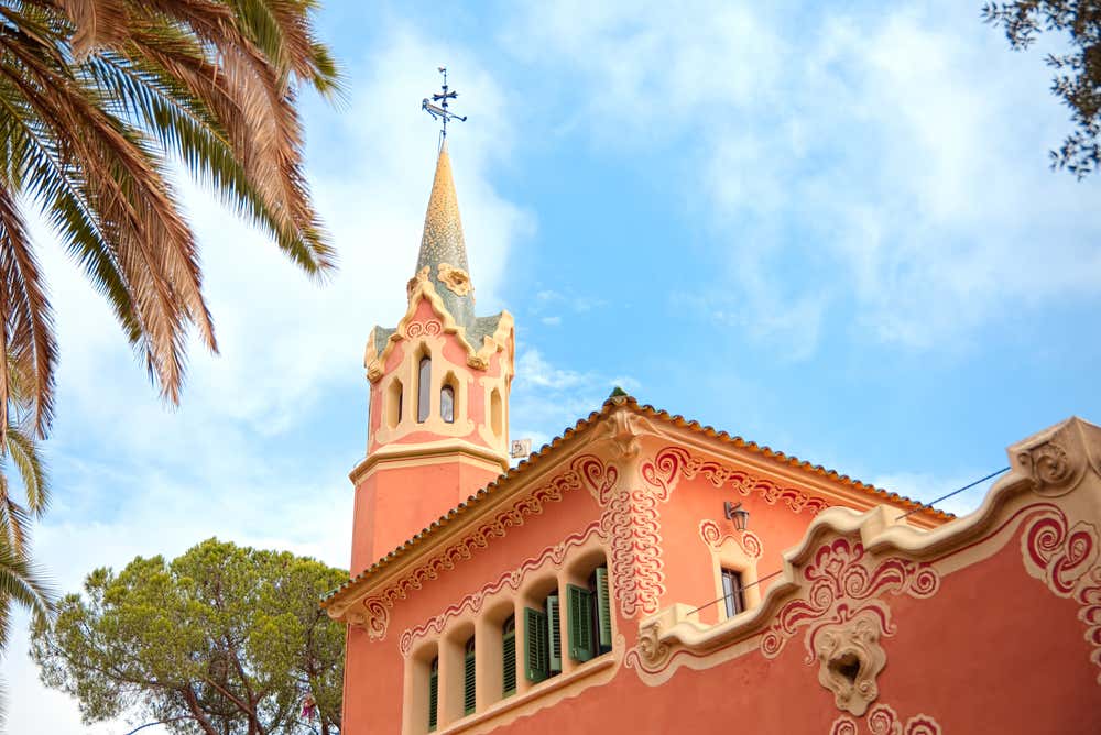La Casa Museo Gaudí al Parc Güell di Barcellona: un edificio rosa con decorazioni in rilievo color crema, persiane verdi e una torretta con la guglia a punta e una croce in ferro battuto. In primo piano si vedono foglie di palma e pini, sotto un cielo azzurro con qualche nuvola