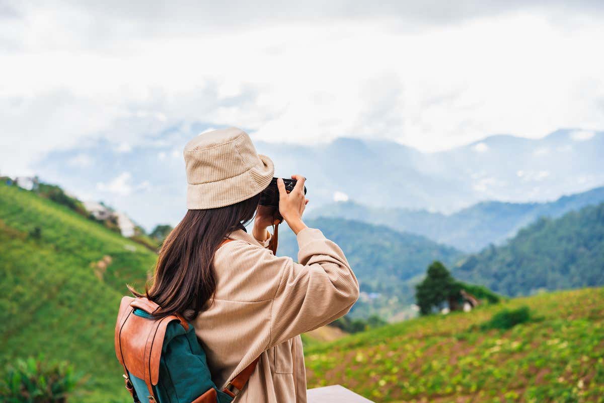 Una viajera con mochila y sombrero de pana toma una foto de las colinas verdes y montañas neblinosas al fondo