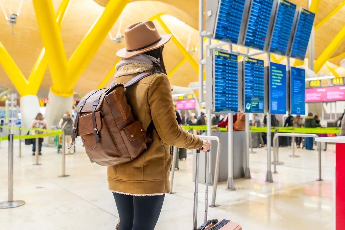 Una mujer con mochila de cuero y maleta observa los paneles informativos de salidas en la terminal