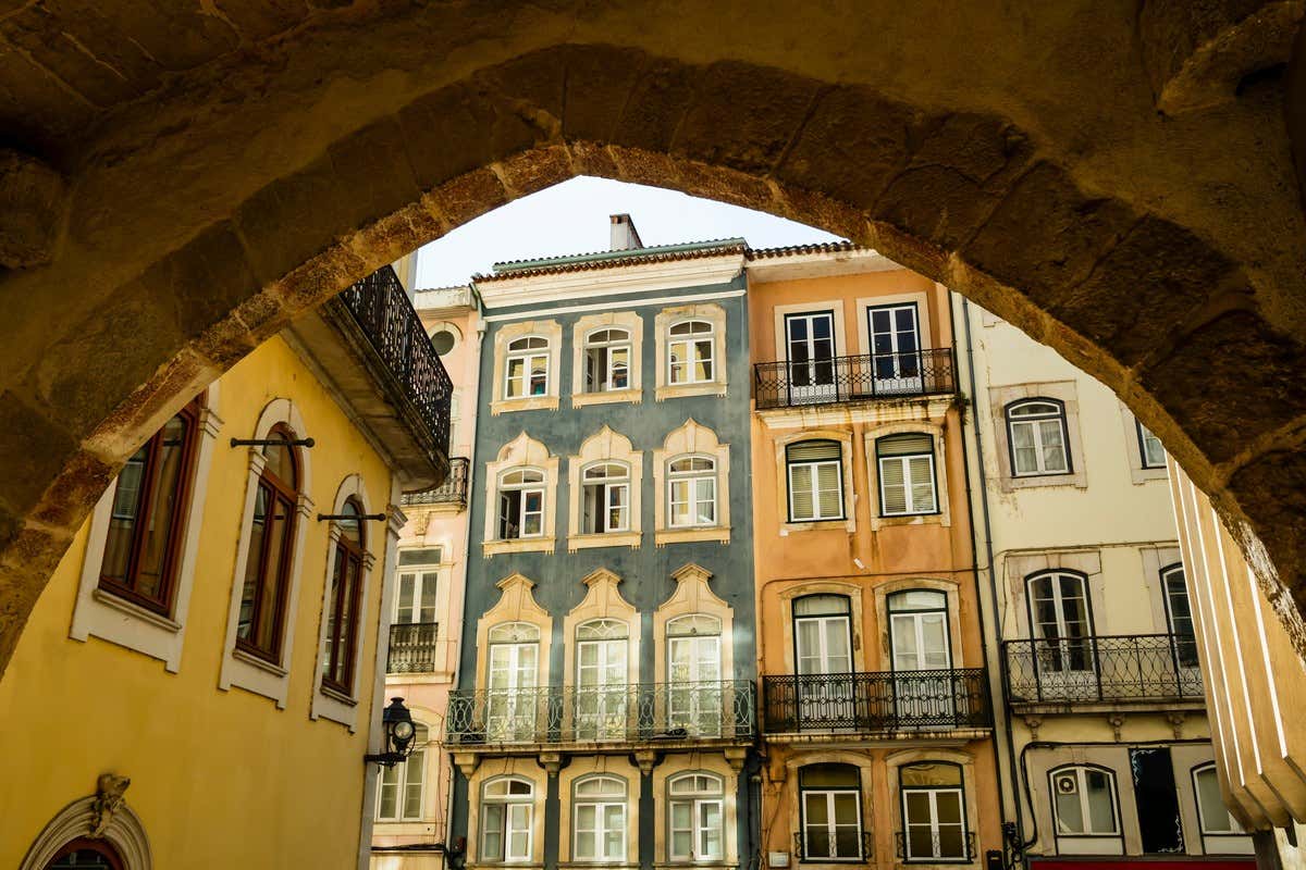 Fachadas coloridas com sacadas de ferro vistas através de um arco de pedra no centro histórico de Coimbra