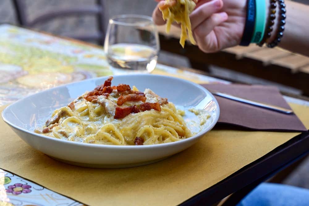 Un piatto di spaghetti alla carbonara serviti all'aperto in un tavolino di un'osteria romana, apparecchiato con tovagliette di carta . La pasta è accompagnata da un calice di vino bianco. Una mano di una persona raccoglie la pasta con un forchetta.