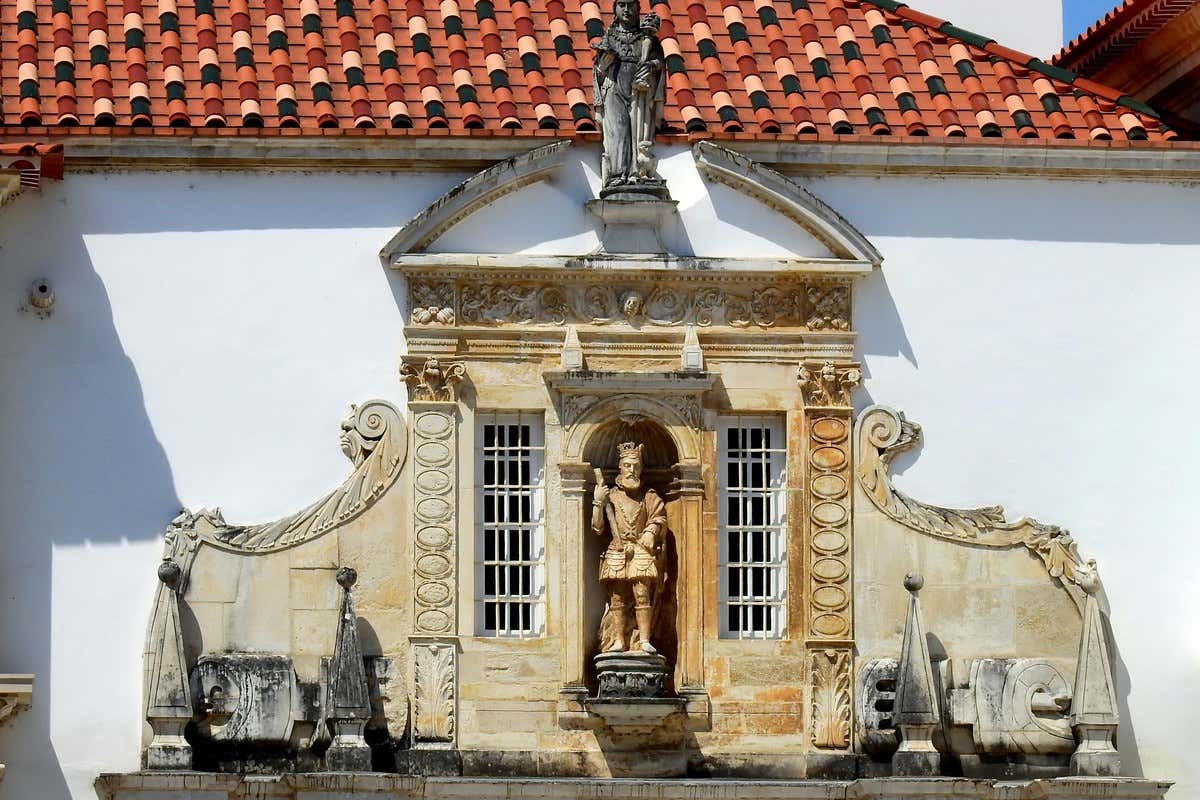 Fachada branca com estátua de um rei em um nicho e telhado de telhas vermelhas na Universidade de Coimbra