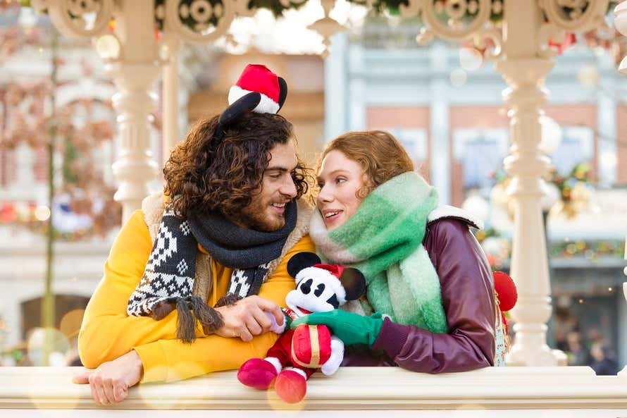 Casal com acessórios festivos, abraçados e segurando um pelúcia do Mickey Mouse natalino