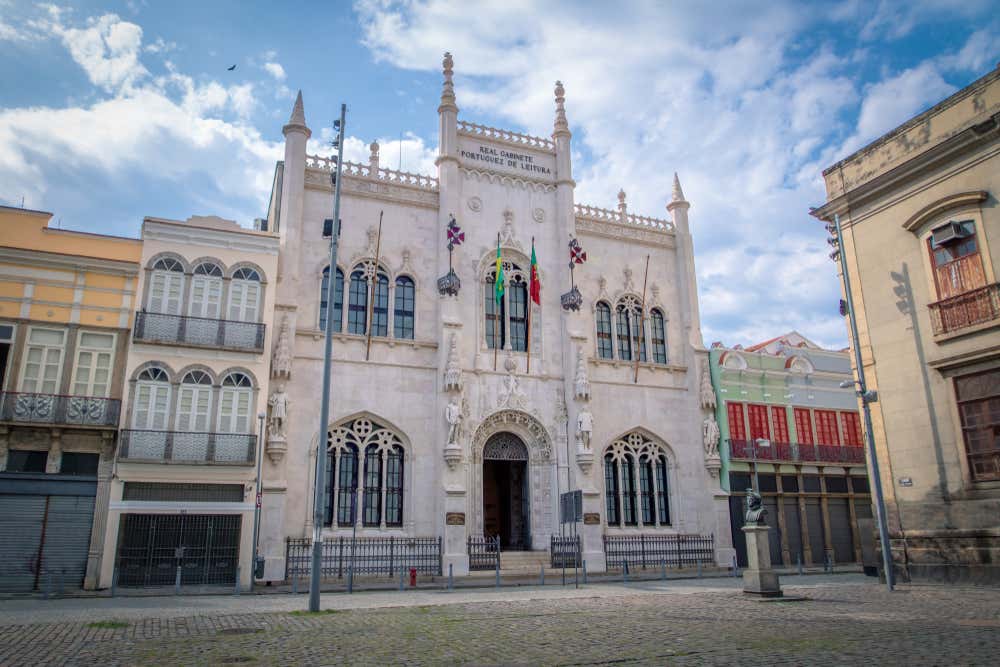 La facciata bianca del Real Gabinete Português de Leitura, nel centro di Rio de Janeiro, in stile neomanuelino. Ha guglie, finestre ad arco gotico, statue scolpite e bandiere che sventolano sopra l’ingresso. Ai lati si vedono palazzi storici con balconi in ferro battuto, sotto un cielo azzurro con nuvole bianche