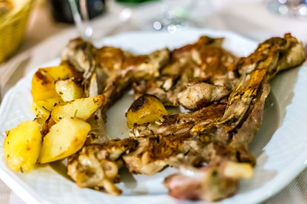 Un piatto bianco con costolette di abbacchio alla scottadito, dorate e croccanti, accompagnate da patate arrosto. Sullo sfondo, sfocati, si vedono bicchieri e bottiglie sulla tavola