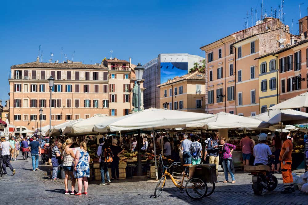 Il mercato di Campo de’ Fiori a Roma, con i banchi di frutta e verdura sotto grandi ombrelloni bianchi e molti passanti tra le bancarelle. Tutto intorno si vedono palazzi storici color ocra e rosa e, al centro, la statua di Giordano Bruno, sotto un cielo azzurro