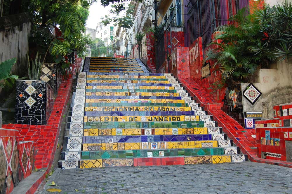 La Scalinata Selarón, a Rio de Janeiro, con i gradini rivestiti di piastrelle gialle, verdi e blu, i colori della bandiera brasiliana, e le scritte “Escadaria Selarón” e “Rio de Janeiro”. Le pareti laterali sono coperte di mosaici rossi e piastrelle decorate, tra piante tropicali e vecchi palazzi del quartiere