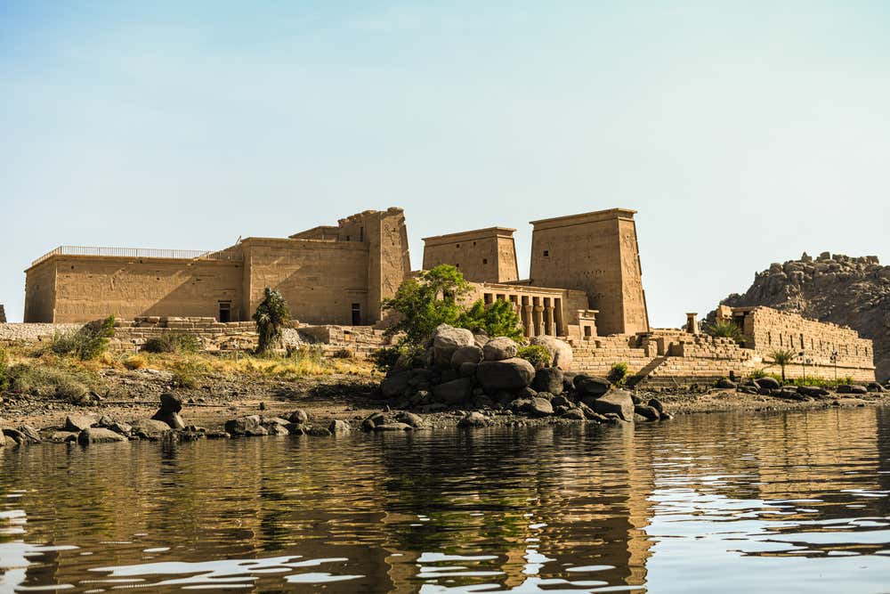 Il tempio di File, sull'isola di Agilkia ad Assuan, visto dall'acqua. I piloni e le mura in arenaria sorgono su una riva rocciosa con qualche palma e cespuglio. In primo piano il Nilo riflette gli edifici, sotto un cielo limpido