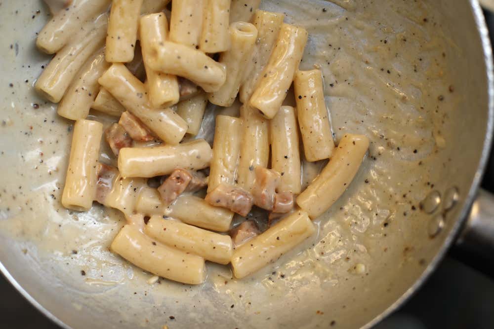Una padella con rigatoni alla gricia mantecati in una crema di pecorino, con pezzetti di guanciale e pepe nero macinato, vista dall’alto