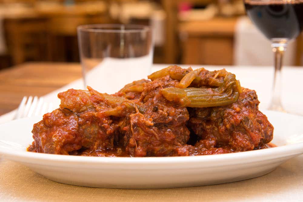 Un piatto di coda alla vaccinara con pezzi di carne di bue cotti a lungo nel sugo di pomodoro e coste di sedano stufato, servito su un piatto bianco. Sullo sfondo, sfocati, si vedono un bicchiere d’acqua, un calice di vino rosso e i tavoli di una trattoria